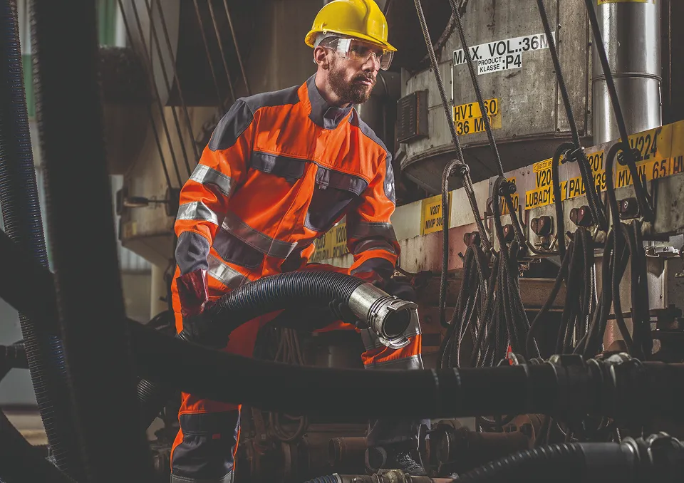 Une personne portant un EPI de couleur orange avec des bandes réfléchissantes argenté, casque jaune et lunette de protection, ainsi que des gants.